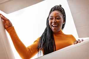 Woman in an orange long sleeve turtleneck with long black, braided hair opening a package and smiling.